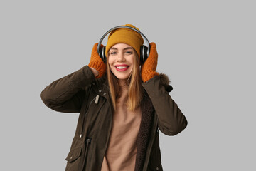 Beautiful young woman in warm winter clothes with headphones on grey background
