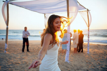 A joyful woman in a white dress dances on the sandy beach during a wedding ceremony as the sun sets over the ocean, surrounded by happy guests. Generative AI.