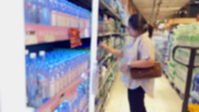 Woman shopper compares bottled water on supermarket shelves, reading a label in the beverage aisle. Bright indoor retail scene with price tags, stacked bottles and product displays