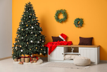 Decorated Christmas tree with gift boxes, sideboard with cushions, Santa hat and sack near yellow wall