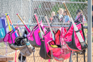 Brightly colored softball bags hang on a chain link fence at a sports field during a practice session in the afternoon sunlight
