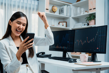 Successful young Asian businesswoman earning high profit raising fist up, looking smartphone for exchange stock market's financial technology with dynamic business graph at modern office. Stratagem.