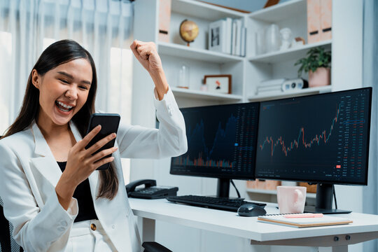 Successful young Asian businesswoman earning high profit raising fist up, looking smartphone for exchange stock market's financial technology with dynamic business graph at modern office. Stratagem.