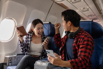 Excited young Asian couple sitting on an airplane, holding a passport, cash, and smartphone while celebrating together. The woman looks happy, and the man shows a cheering gesture, enjoying the moment