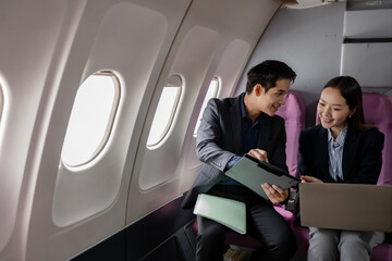 Asian business professionals working together on a laptop while sitting on a commercial airplane. They appear to be discussing a project or presentation in a productive and collaborative manner.