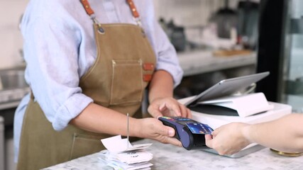 Senior entrepreneur cafe owner barista holds POS terminal while customer swipes creditcard, Asian retired female in apron smiling at counter, demonstrating cashless payment inside modern coffee shop - Powered by Adobe