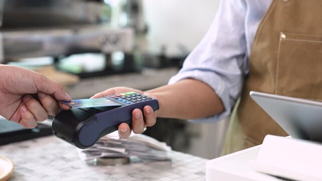 Senior entrepreneur cafe owner barista holds POS terminal while customer swipes creditcard, Asian retired female in apron smiling at counter, demonstrating cashless payment inside modern coffee shop
