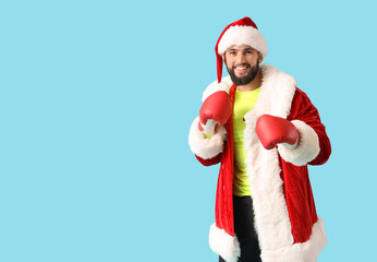 Young happy sporty man in Santa costume with new boxing gloves as Christmas gift on blue background