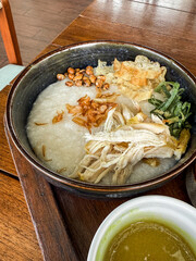 Traditional Indonesian chicken porridge or Bubur Ayam served with crackers, chili sauce, green sauce, and toppings on a wooden tray over rustic wooden table.