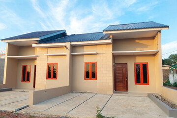 A simple permanent house with a modern minimalist design, beige walls, wooden doors, and red-framed windows, viewed from the front under a clear blue sky.