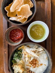 Traditional Indonesian chicken porridge or Bubur Ayam served with crackers, chili sauce, green sauce, and toppings on a wooden tray over rustic wooden table.