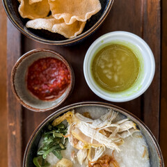 Traditional Indonesian chicken porridge or Bubur Ayam served with crackers, chili sauce, green sauce, and toppings on a wooden tray over rustic wooden table.