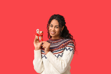 Beautiful young African-American woman in stylish earrings with gift box and candy cane on red background. Christmas celebration