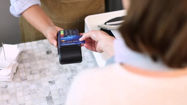 Senior entrepreneur cafe owner barista processes credit card payment on terminal, Asian female in apron smiling toward client across tiled counter, secure cashless sales inside modern coffee shop