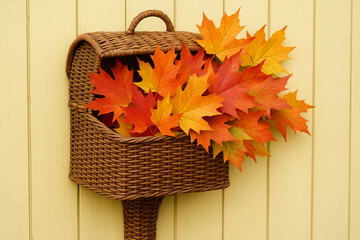Wicker mailbox overflowing with vibrant autumn leaves, showcasing shades of orange and yellow against soft yellow background, evokes warm, seasonal feeling