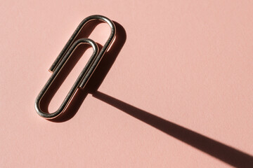 Metallic paperclip rests on soft pink background, casting long shadow that adds depth to simple composition. contrast between shiny metal and pastel surface creates visually appealing scene