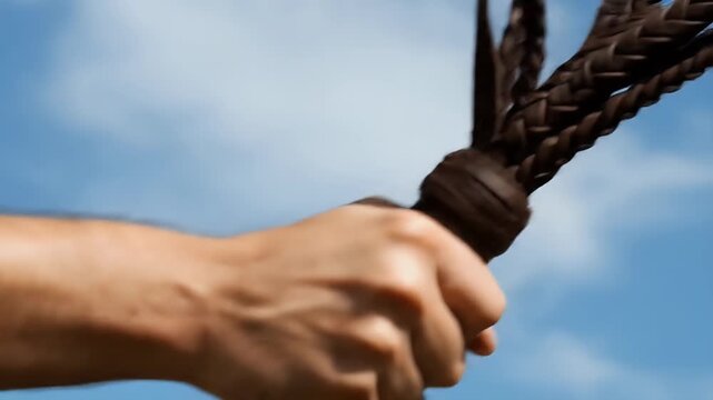 Close up of a hand holding a braided leather whip against a blue sky.