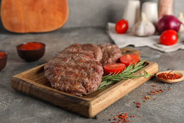 Wooden board with tasty meat cutlets, tomato, rosemary and paprika on grey grunge background