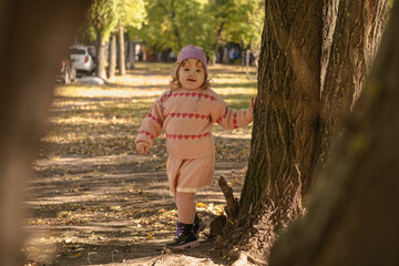 A young child with curly hair runs joyfully through a park covered in colorful autumn leaves. She wears a cozy sweater and a cute hat, enjoying the beautiful sunny day.