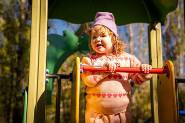A young child enjoys playing on a playground in a lovely park. Dressed warmly, the child smiles while exploring the climbing structure under a clear blue sky.