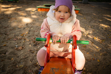 Child plays on a red playground toy in a sunny park surrounded by trees and benches
