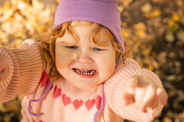 A young child with curly hair and a purple hat seems upset while reaching out with both hands. The park is filled with fallen leaves, showcasing the vibrant colors of autumn on a sunny day.