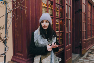 A young girl tourist in an autumn coat, a wide scarf and purple angora hat with a smartphone stands alone,leaning against the wooden wall. The concept of a navigator,social networks,internet,messenger