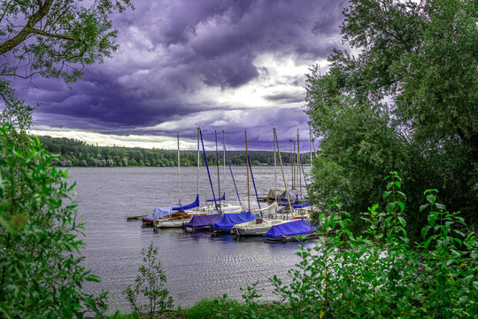 Eine malerische Seelandschaft, dramatisch eingerahmt von leuchtend gr&uuml;nem Laub. Im Zentrum liegen kleine Segelboote vert&auml;ut, deren Masten in einen Himmel voller massiver, dunkellila Wolken ragen. Die 