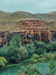 Vertical photo of a river flowing near black rocky formations with rolling hills in the background under an overcast sky.