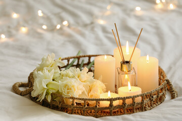 Tray with reed diffuser, burning candles and flowers on bed in bedroom, closeup