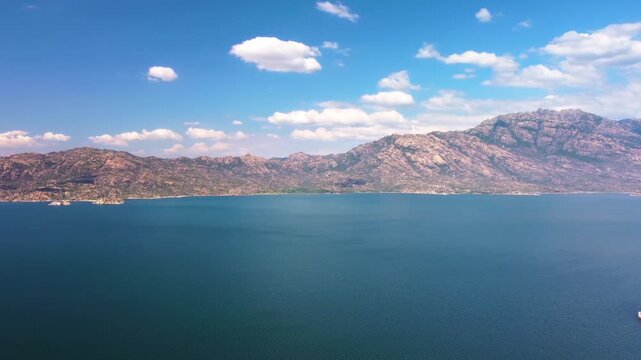 Drone footage captures the tranquil beauty of Bafa Lake in Turkey, showcasing its vast expanse of blue water, surrounded by rugged mountains under a clear, bright sky with scattered clouds. Ideal for 