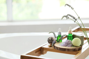 Wooden bathtub tray with bath bombs, lavender flowers and bottles of cosmetic products in  bathroom, closeup