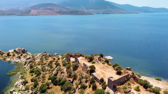 An aerial view captures the serene blue waters of Lake Bafa, Turkey, with a rocky peninsula covered in ancient ruins and olive trees extending into the lake.