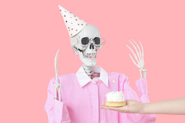 Human skeleton in party hat and female hand with birthday cake on pink background, closeup