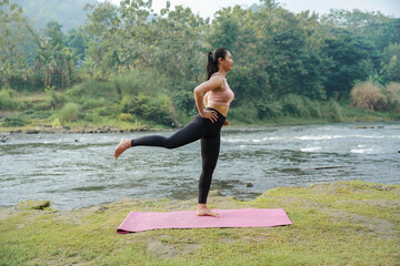 Fototapeta premium A slender Asian girl with brown skin in sportswear doing stretching movements and swinging her legs in the open air on the edge of a beautiful river, before doing sports.