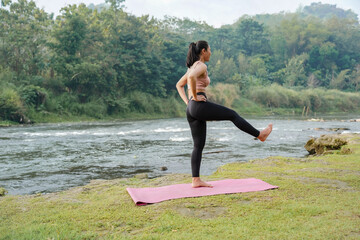 Fototapeta premium A slender Asian girl with brown skin in sportswear doing stretching movements and swinging her legs in the open air on the edge of a beautiful river, before doing sports.
