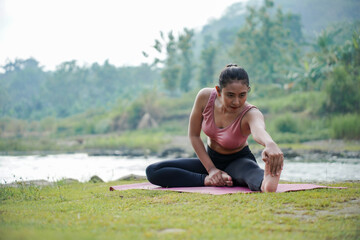 A slender Asian girl with brown skin in sportswear is doing stretching movements of her hamstrings in the open air on the edge of a beautiful river, before doing sports.