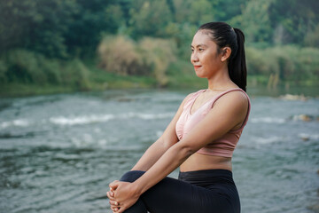 A slender Asian girl with brown skin in sportswear doing muscle warm-up movements in the open air on the edge of a beautiful river, before doing sports.
