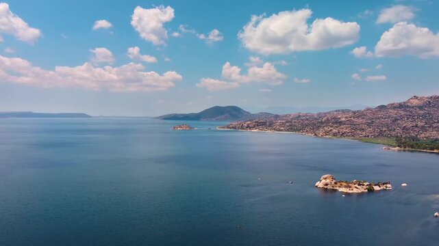 Aerial footage capturing the tranquil beauty of Bafa Lake in Turkey, showcasing its vast blue waters, surrounding mountains, and scattered islands under a clear sky with fluffy white clouds.