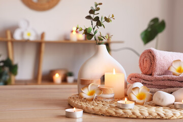 Wicker mat, plumeria flowers, spa stones, burning candles and jars of cosmetic products on wooden table in spa salon, closeup