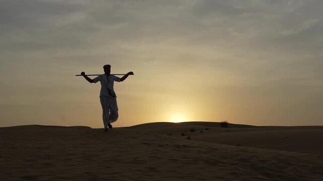 Silhouette of a Rajasthani man walking on a sand dune in the Thar Desert near Jaisalmer, Rajasthan, India. Sunset, with the sky and the sun's glow creating a dramatic backdrop