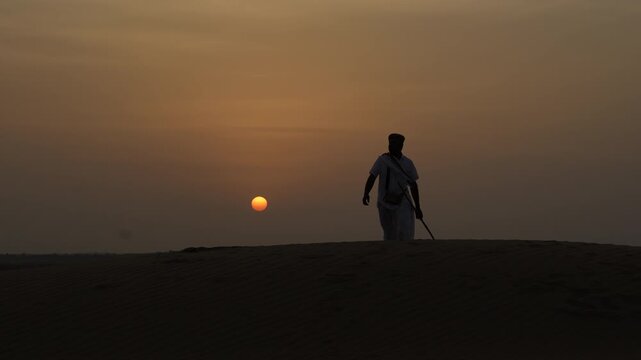 Silhouette of a Rajasthani man walking on a sand dune in the Thar Desert near Jaisalmer, Rajasthan, India. Sunset, with the sky and the sun's glow creating a dramatic backdrop