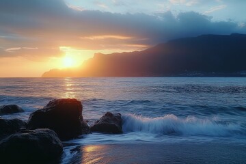 Sunset over the ocean with gentle waves crashing on dark rocks and silhouetted mountains under a partly cloudy sky