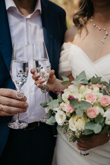 The bride in a white dress holds a bouquet of multi-colored roses in one hand and a glass of champagne in the other, next to another glass held by the groom in a classic suit