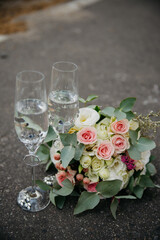 Two glasses of sparkling white wine stand on the asphalt next to a bouquet of colorful roses on their wedding day