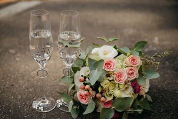 Two glasses of champagne stand on the asphalt next to a bouquet of colorful roses on a wedding day in the middle of a pine forest