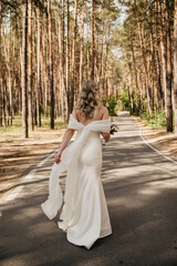 A young slender bride in a white tight dress holding a bouquet of flowers is walking along an asphalt road in a pine forest