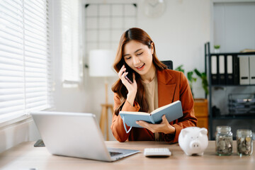 Asian businesswoman talking on smartphone and writing in notebook at office desk with piggy bank...