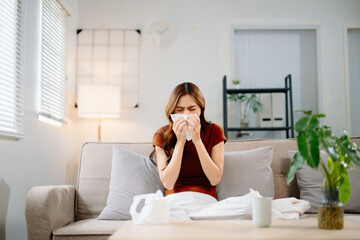 Young Asian woman suffering from flu symptoms, covered with a blanket on a sofa. Concept of illness, healthcare, and recovery. Stay indoors, stay healthy