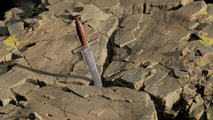 A long, sleek sword stands upright within a rocky crevice, illuminated by soft twilight glow. The surrounding stones display textures and shades, evoking a sense of mystery and history. © icetray